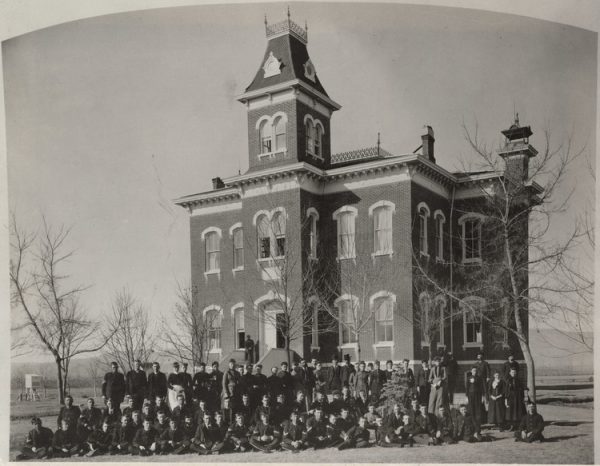 A group of young men and women stand, kneel, sit, or lounge in front Old Main in 1886. 