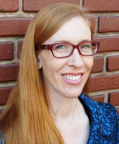 Dr. Julia Schleck. A woman with long, red hair and glasses wearing a blue collared shirt against a brick building.