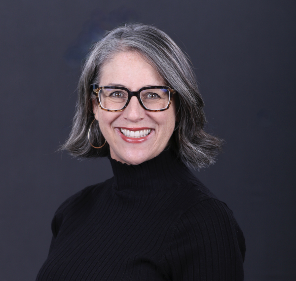 Professional headshot of a woman with grey chin-length haircut and glasses wearing a black turtleneck sweater.