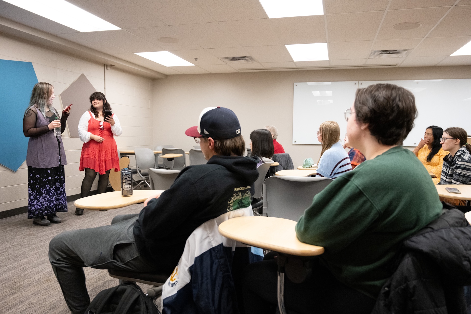 Two students presenting to a classroom of students seated at tablet-arm desks. One presenter wears a gray outfit, the other a red dress. Students are facing forward, listening to the presentation.