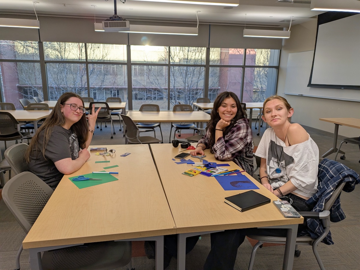Three students posing together at a table with art supplies and blue paper materials spread out.