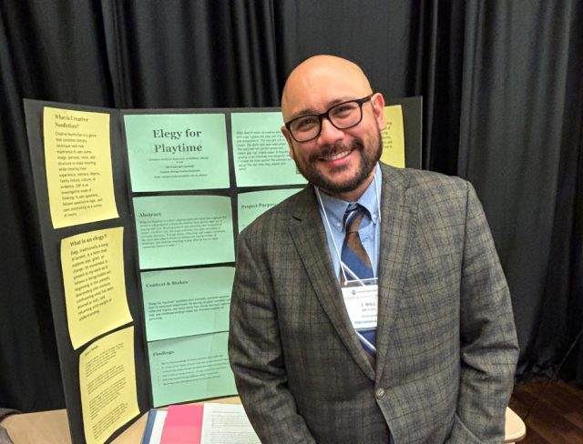 GradShow_web10 Will Brooks in checkered blazer standing beside poster about "Elegy for Playtime" at Graduate Student Showcase, Nov. 19.