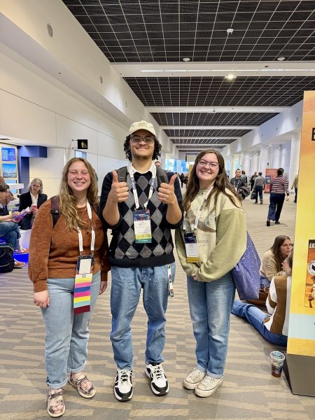 Three students smile and give thumbs up in a convention center hallway with solar panel ceiling visible.
