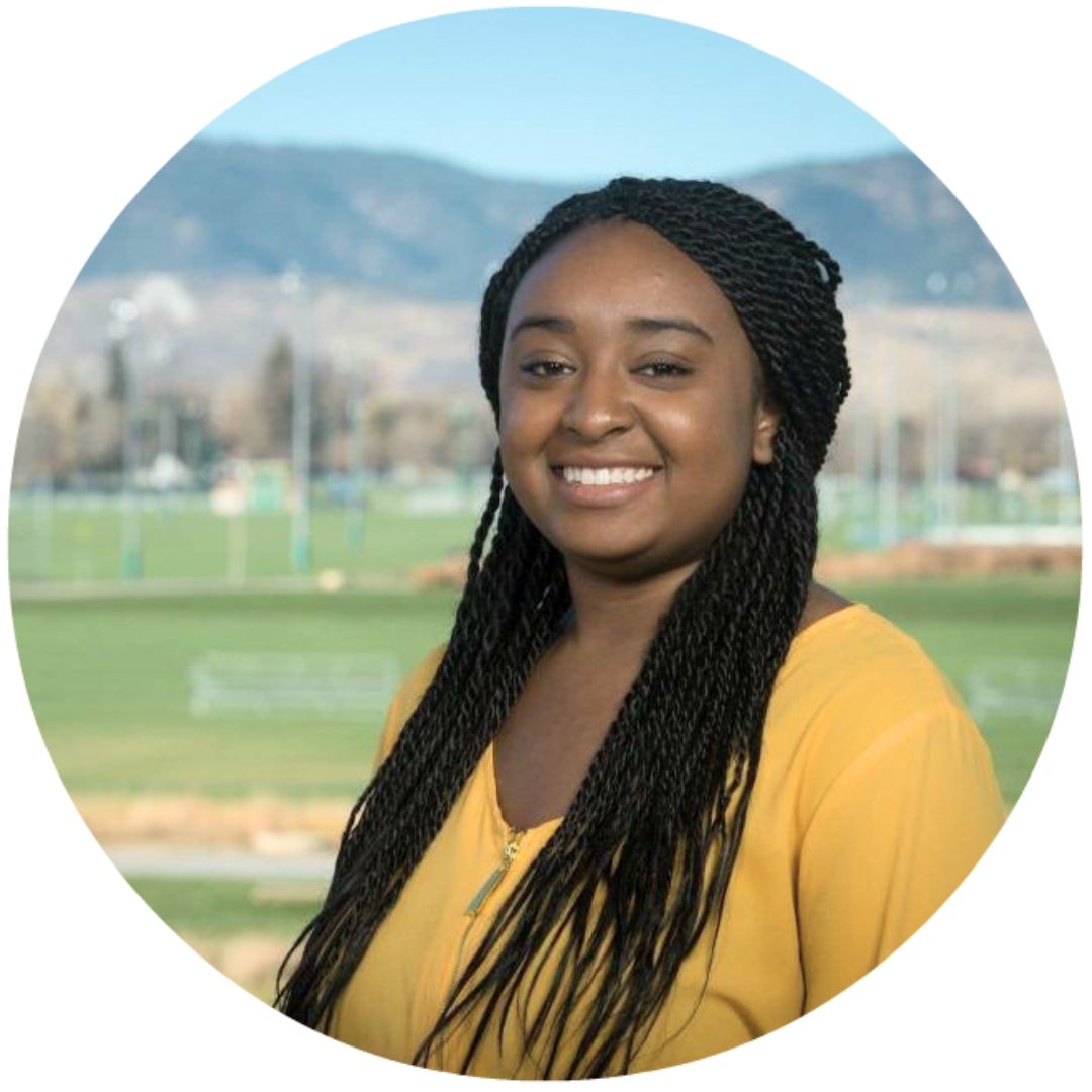 A person in a bright yellow top smiles warmly at the camera in a professional headshot-style photo. They have long black braids and are photographed against a blurred background showing fields and mountains.