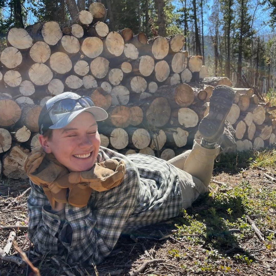 Max Pohl smiles, lying on the ground next to a large stack of cut logs in a forest setting in a plaid shirt and cap.