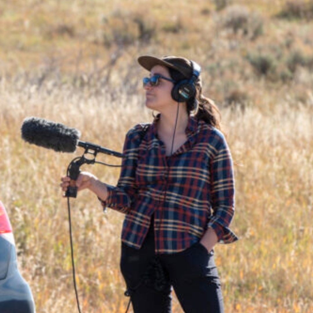 A person wearing a plaid flannel shirt, baseball cap, sunglasses and headphones operates audio recording equipment with a boom microphone in an outdoor setting with golden grass in the background.