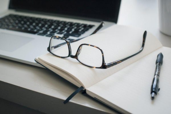 A pair of glasses and a pen on an open notebook next to a laptop