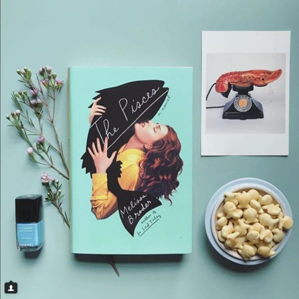A book called "The Pisces" surrounded by goldfish, nail polish, dried flowers, and Polaroids.