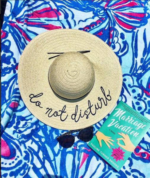 A hat that says "do not disturb" laid on a beach towel with a book called "Marriage Vacation." 