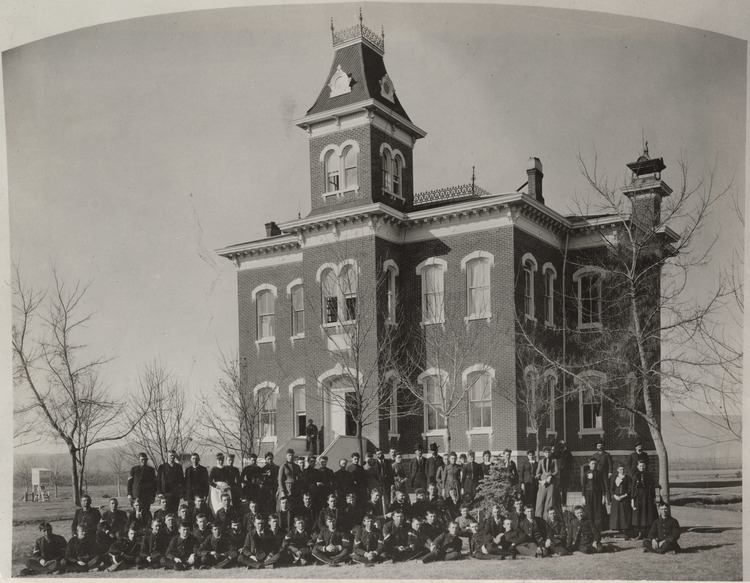 A group of young men and women stand, kneel, sit, or lounge in front Old Main in 1886.