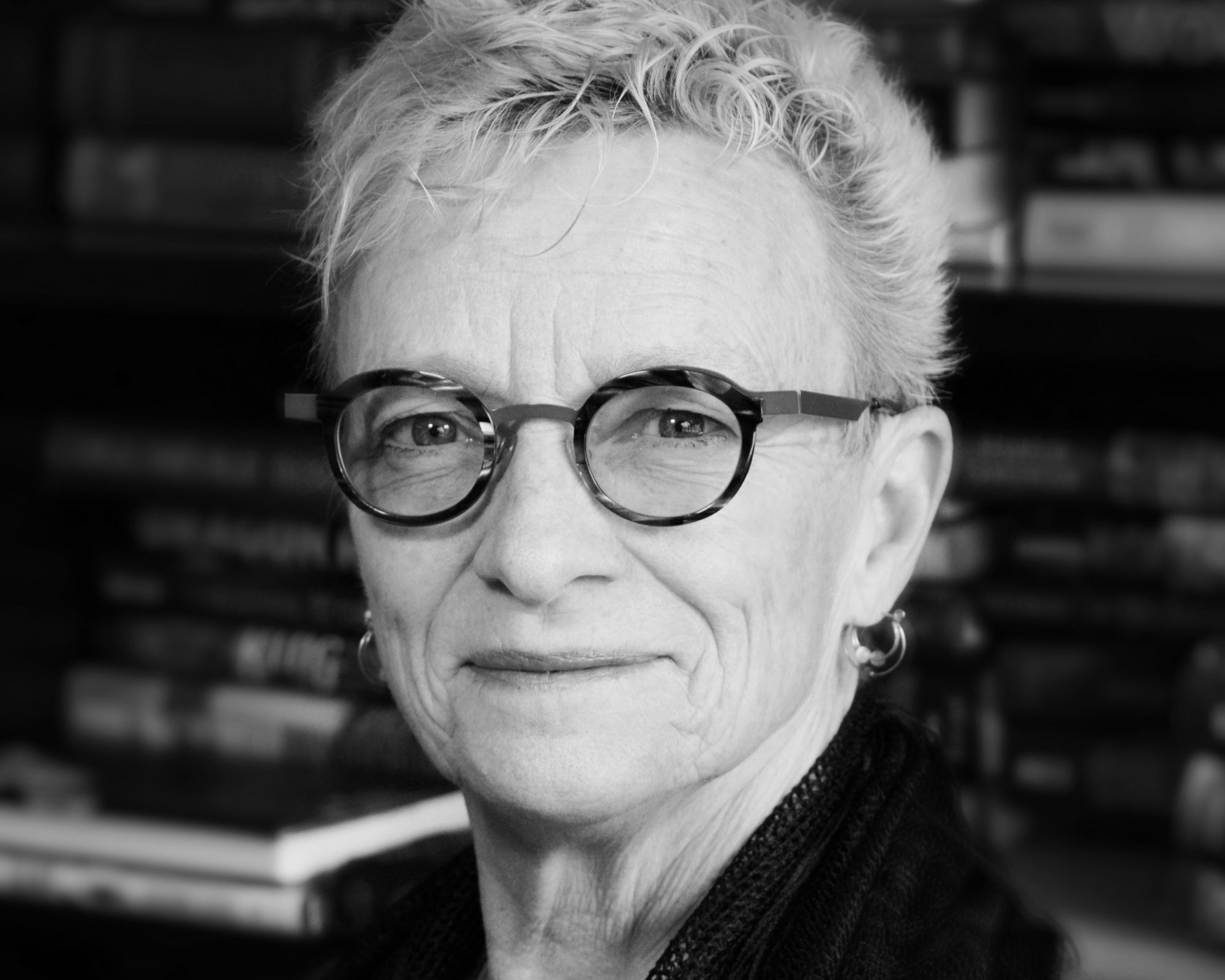 A black and white photo of Beth Lechleitner looking at the camera with stacks of books in the background