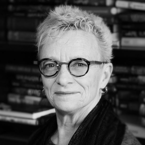 A black and white photo of Beth Lechleitner looking at the camera with stacks of books in the background