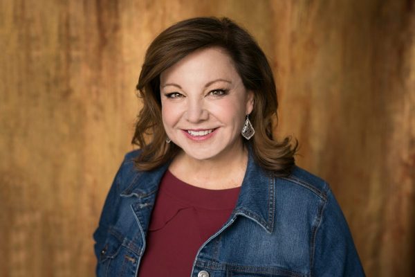 Donna Cooner in a jean jacket over a red shirt in front of a wood background