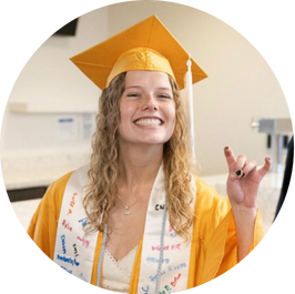 Annie Ness smiles and makes 'ram horns' gesture at commencement ceremony. She wears a gold cap and gown and white stole.