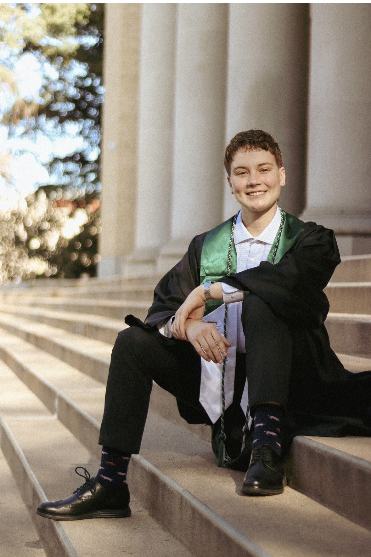 Max Pohl, wearing a black graduation gown with a green satin stole and honor cords, smiles while sitting on stone steps in front of a building with large columns.