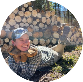 Max Pohl smiles, lying on the ground next to a large stack of cut logs in a forest setting in a plaid shirt and cap.