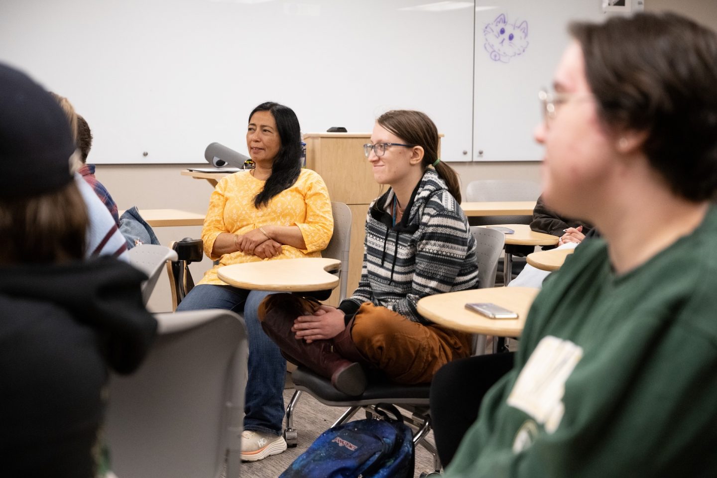 Students sitting in a modern classroom with tablet-arm chairs, listening to a presentation. A professor in a yellow top is visible in the center, with other students in casual clothing around her.