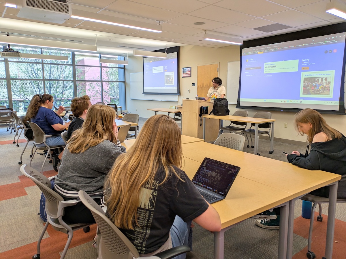 Students attending a presentation in a classroom with large windows, student presenter at front with projected content on screen.
