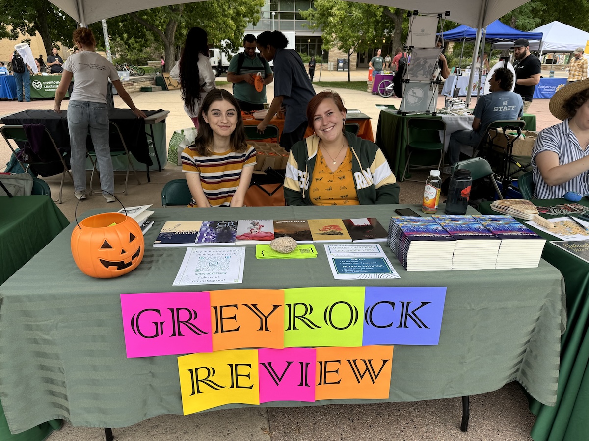 Two students staffing the Greyrock Review booth at an outdoor event, with colorful lettered signs spelling out the publication name and stacks of magazines displayed on the table.
