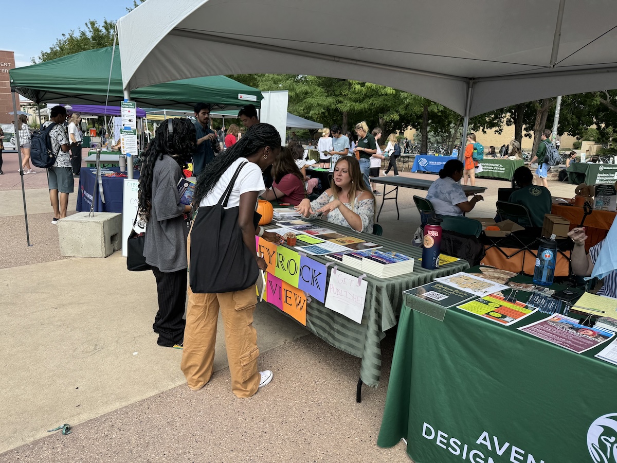 Outdoor campus fair with students visiting a Greyrock Review information booth under white tents, with volunteers distributing materials.