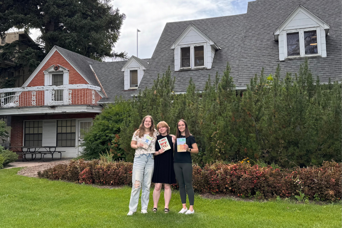 Three graduate students smile and pose together outside campus building.
