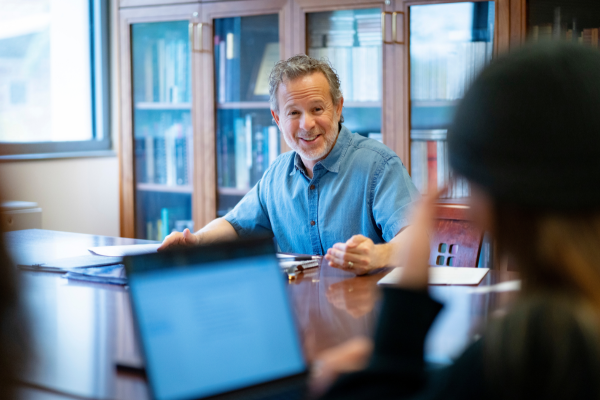 Faculty member smiles in classroom toward student with open laptop.