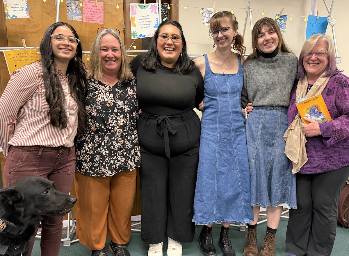 Six smiling student interns and faculty pose together at an event celebrating 20 years of SpeakOut! Journal, in front of a colorful story wall display with signs reading "We are all unfinished stories. What's your next chapter?" The person on the far right holds a copy of a book titled Unfinished Stories. A black Labrador in a service vest is visible at lower left.