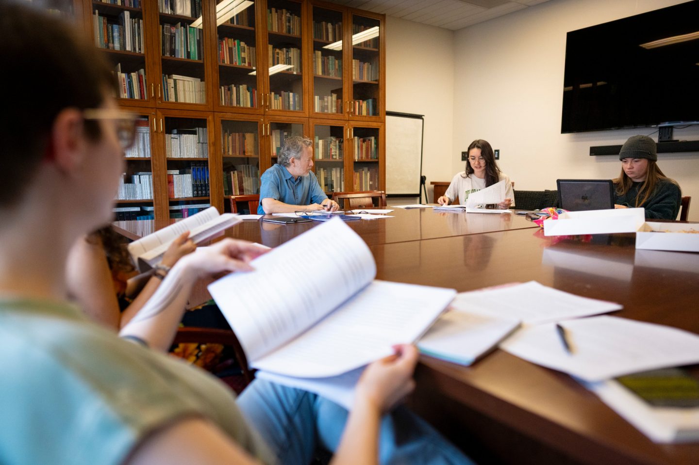 Students sit around a long wooden table in a book-lined library or seminar room, reading and flipping through books and papers, with a professor seated at the far end.