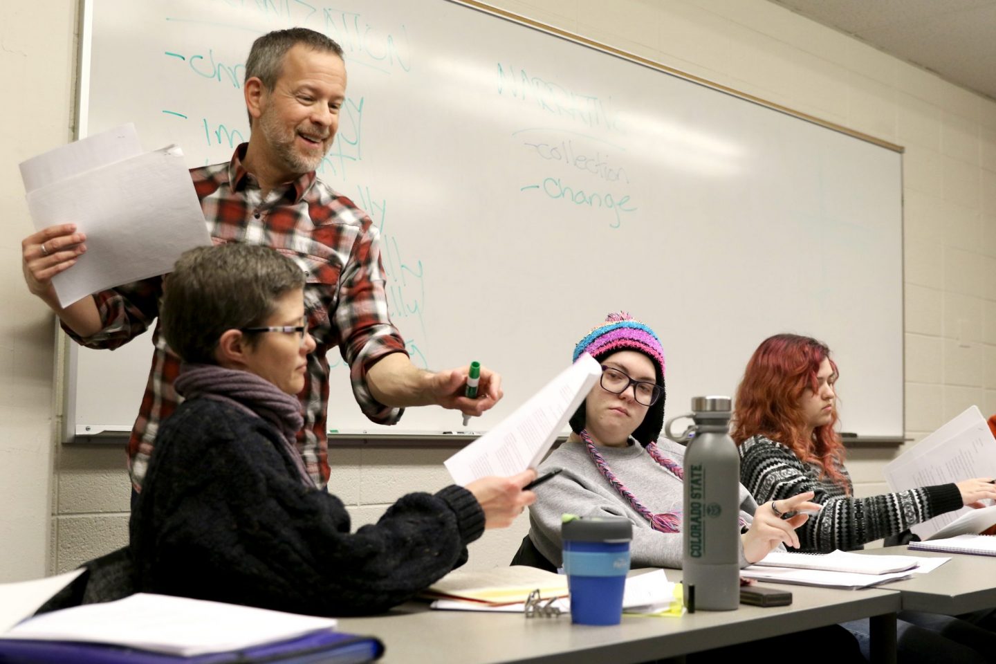 Instructor holding papers and a marker addresses seated students in a classroom, with notes about "Narrative," "collection," and "change" visible on the whiteboard behind him.