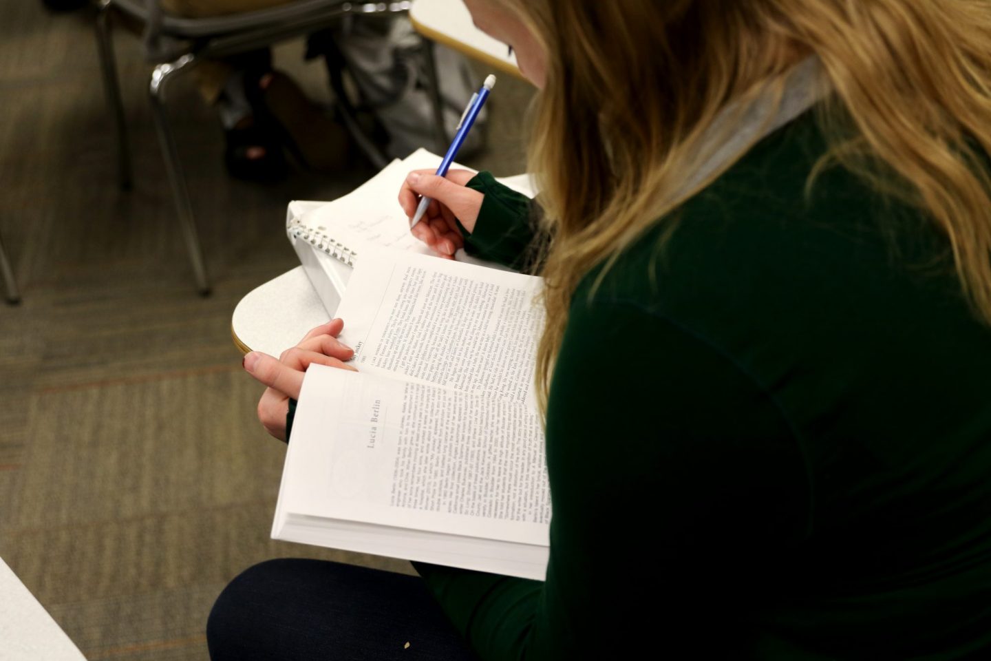 Overhead view of a student in a green sweater taking notes with a pen while reading a book open to a page about author Lucia Berlin.