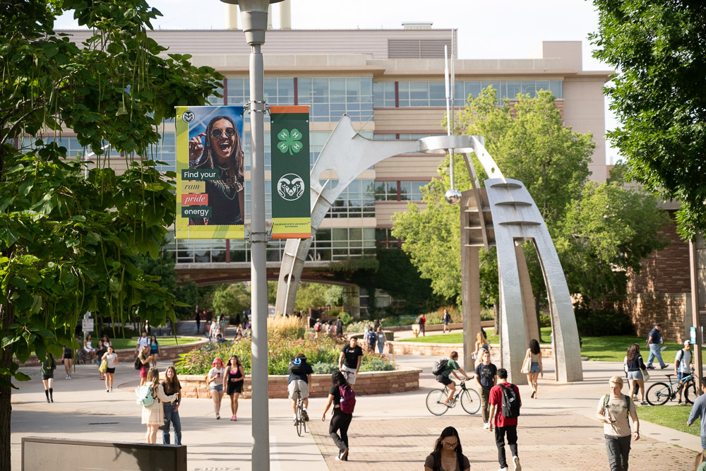 Colorado State University campus plaza with an arched metal sculpture, a "Ram pride energy" banner, and students walking and biking past a large building.