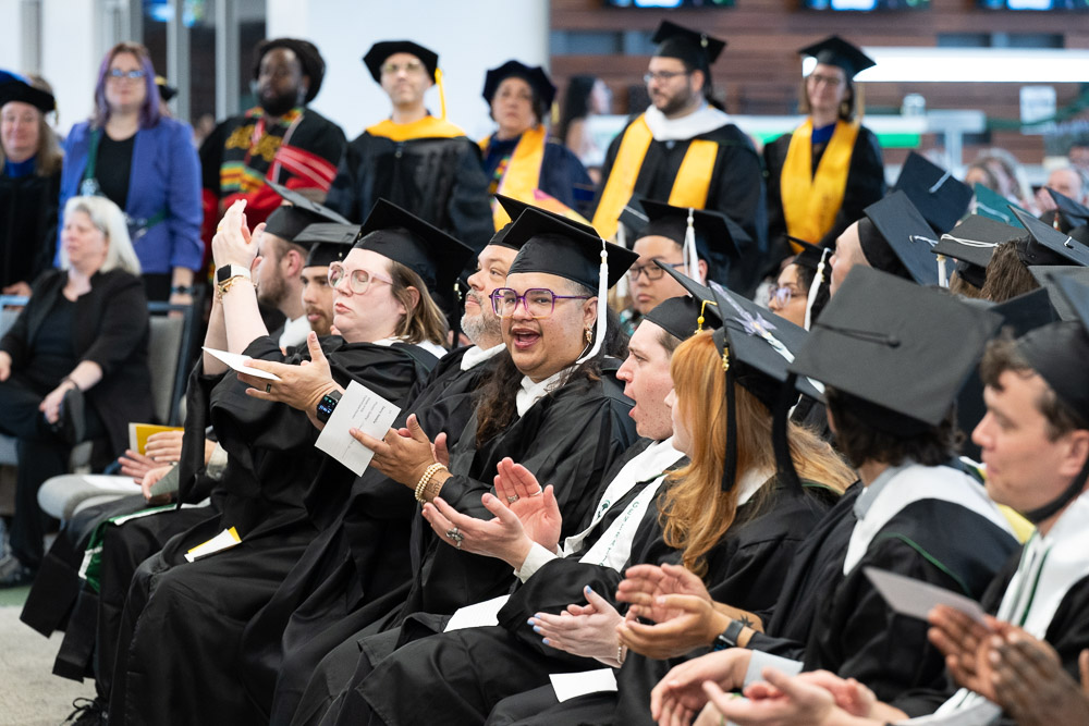 Graduates in caps and gowns clap and cheer during a Colorado State University commencement ceremony, with faculty in colorful academic regalia standing in the background.