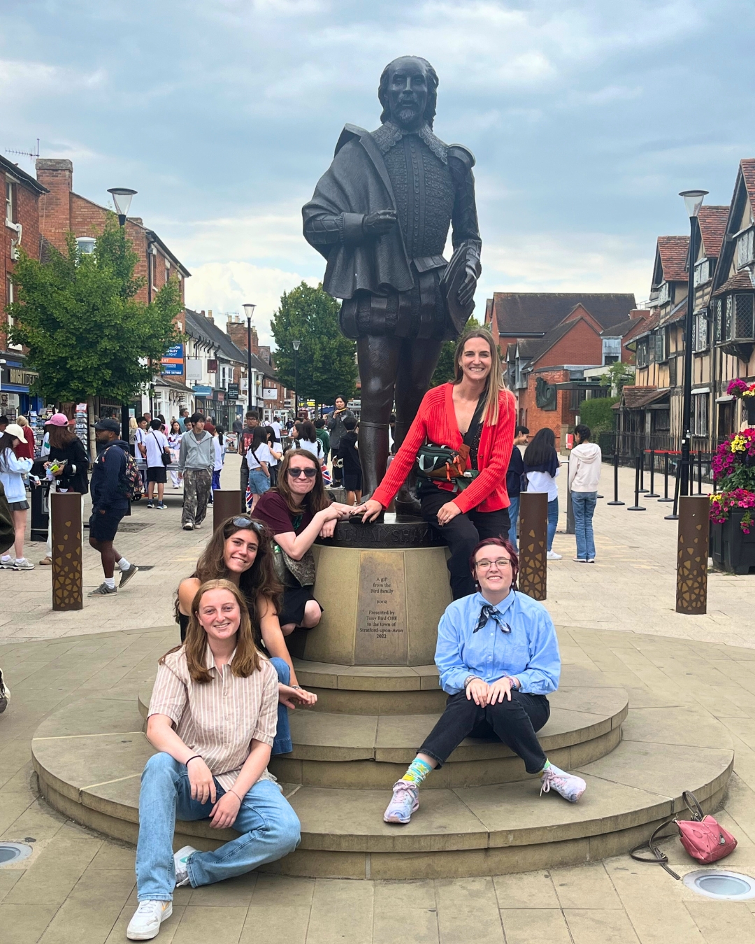 Group posing around the base of a bronze statue on a busy pedestrian street lined with shops.