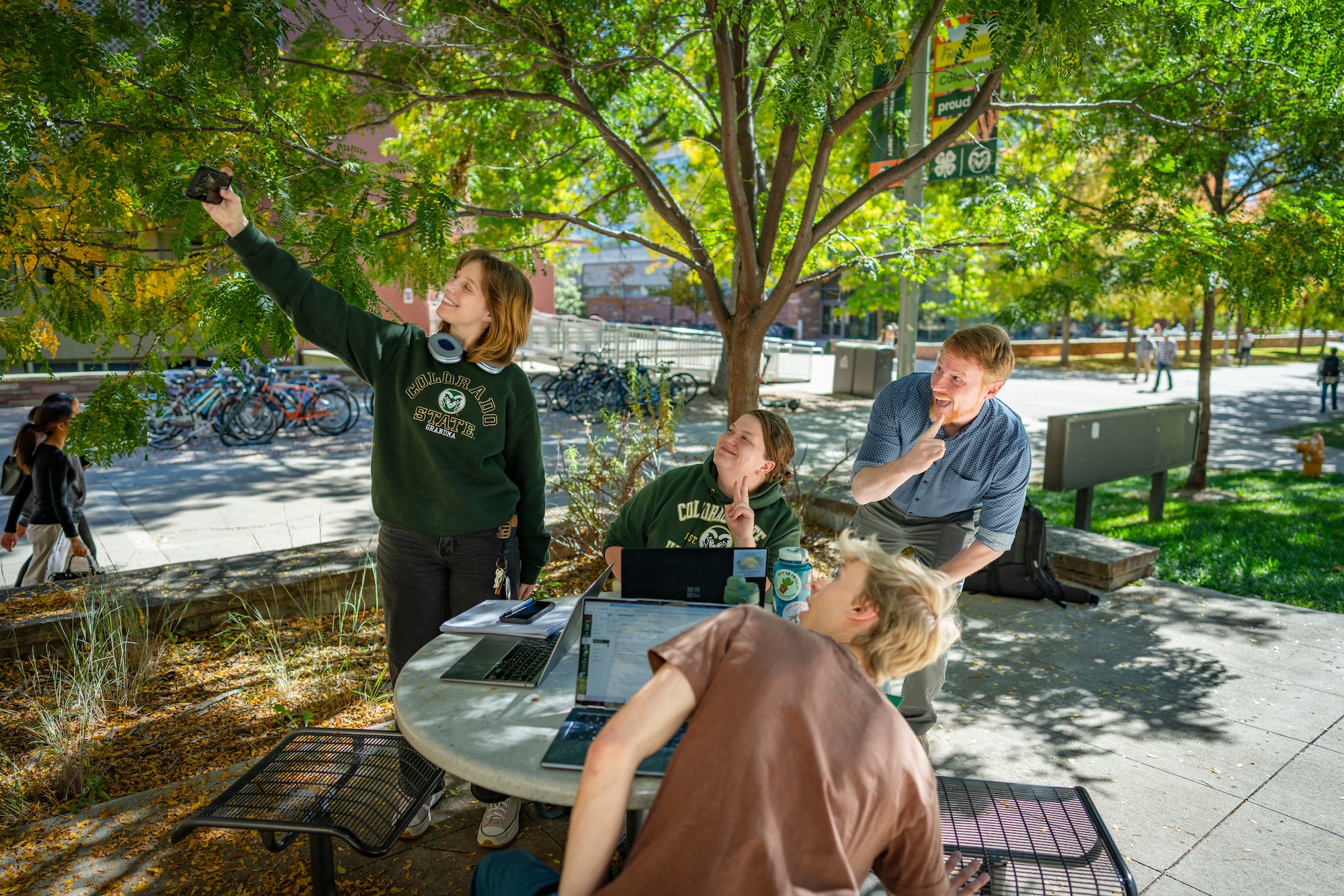 Students and instructor take a group selfie at an outdoor table on a college campus, laptops open in front of them, with bike racks and buildings in the background.