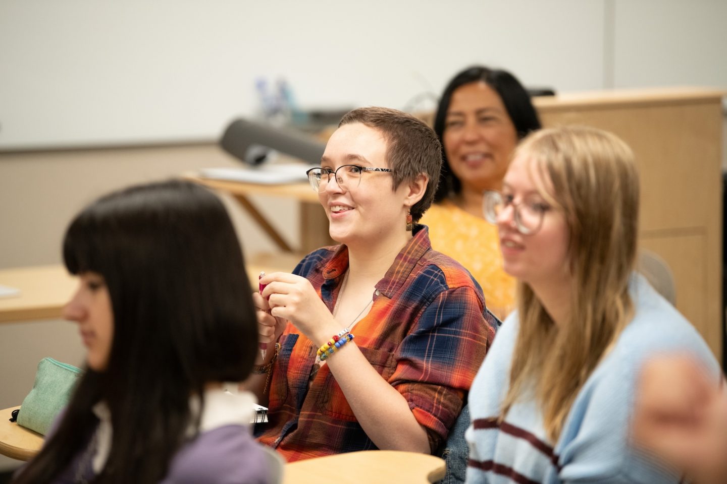 People in a classroom setting, with the person in the foreground wearing glasses and a colorful plaid shirt, smiling while engaged in discussion. Three other people are visible in the background, also appearing to be participating in the conversation.