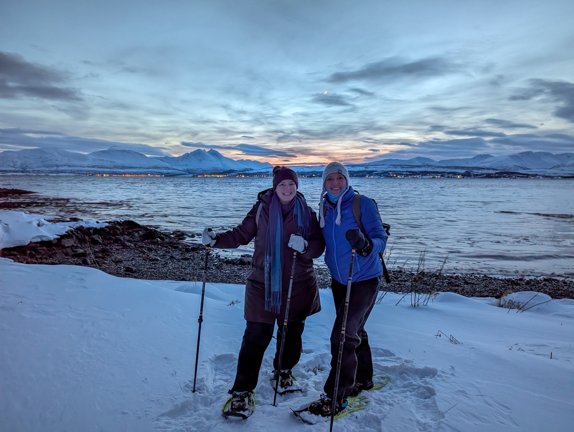 Rosa Martey and Naitnaphit Limlamai in snowshoes and winter gear smile for a photo on a snowy shoreline at dusk, with a fjord, snow-capped mountains, and a colorful sunset sky in the background.
