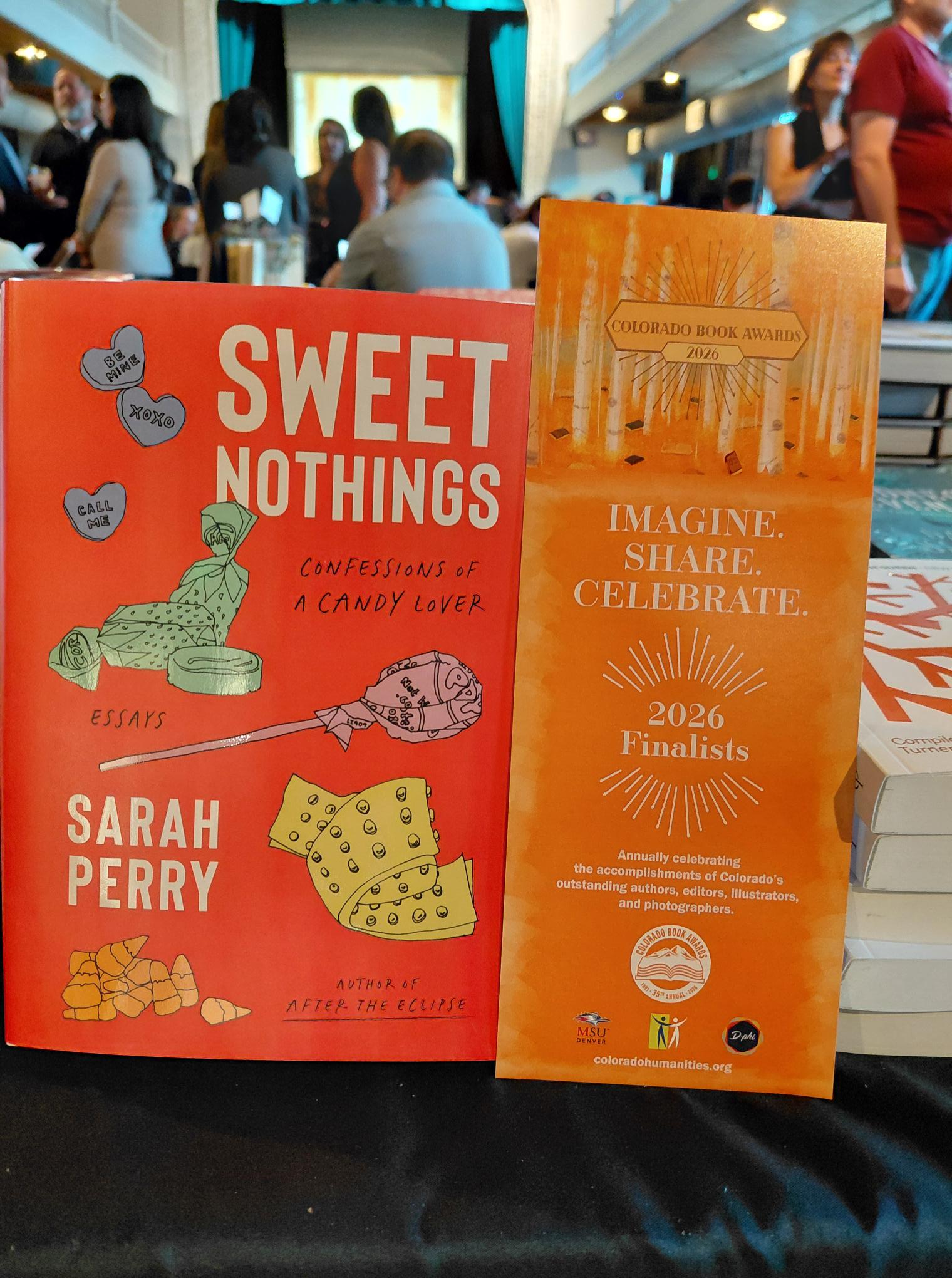 Sarah Perry's book "Sweet Nothings: Confessions of a Candy Lover" displayed next to a sign for the 2026 Colorado Book Awards finalists, with event attendees visible in the background.