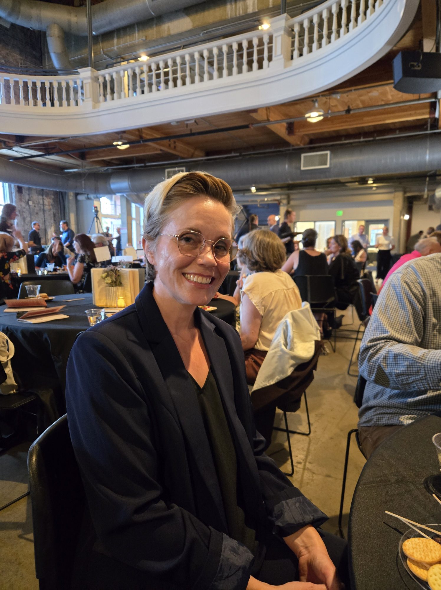 CSU professor Sarah Perry smiles at a table during an indoor event, with other attendees seated and mingling in the background under a high-ceilinged venue with a balcony.