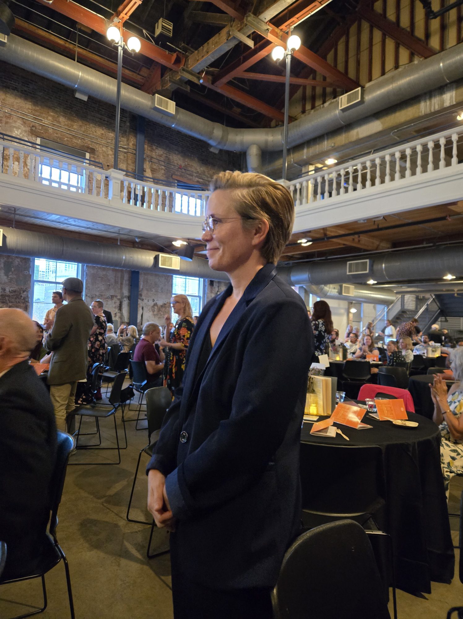 Sarah Perry stands smiling in profile at an indoor event space with brick walls, exposed pipes, and a balcony, guests seated at tables in the background.